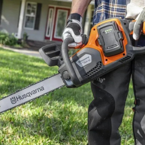 A person holds a Husqvarna chainsaw on a grassy lawn, with a house in the background. They're wearing gloves and rugged work pants, suggesting yard work.