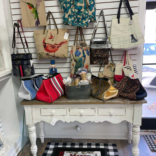 A white wooden table with various colorful bags and clutches displayed. More bags hang on a grid wall above, featuring bird and floral designs. Cozy and artistic vibe.