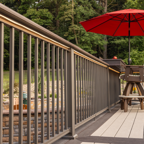 A wooden deck features a sleek metal railing and a vibrant red umbrella beside wooden picnic tables, set against a lush, green forest backdrop.