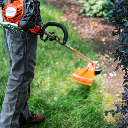 Person using an orange trimmer to cut grass along a garden edge. Wearing jeans and brown shoes, the scene conveys a sense of focus and tidiness.