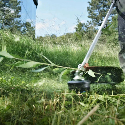 A person uses a string trimmer to cut tall grass in a lush, green field. Background features a blue sky and trees, conveying a peaceful outdoor setting.