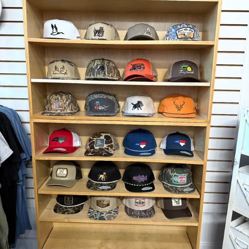 A wooden display shelf with four rows of various trucker hats featuring wildlife and outdoor designs. Colors include camo, orange, and beige. Casual setting.
