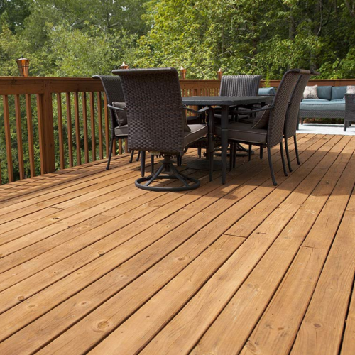 Spacious wooden deck with dark wicker chairs and a black table, surrounded by lush green trees, conveying a serene and inviting outdoor setting.