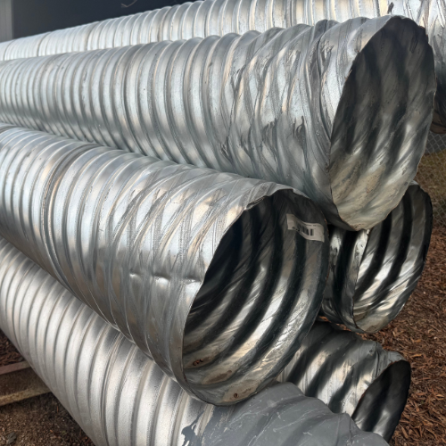 Stack of large, silver corrugated metal pipes on a dirt ground, conveying an industrial tone. The pipes' open ends reveal their hollow interiors.