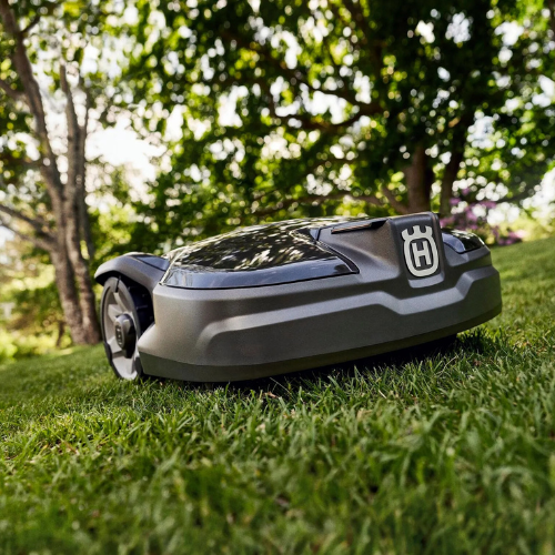 A robotic lawn mower on vibrant green grass under dappled sunlight, with trees in the background. The setting feels serene and high-tech.