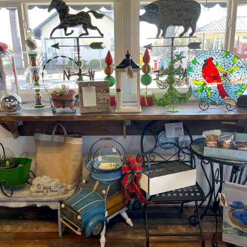 A cozy store display with colorful metal animal sculptures, a stained glass piece featuring a red bird, and a charming blue car planter. Warm, eclectic vibe.