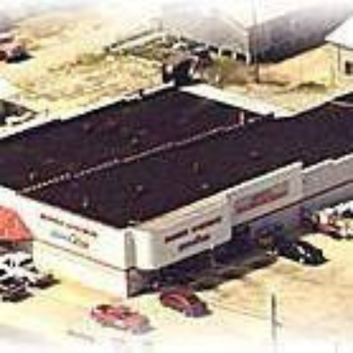 Aerial view of a large car dealership with a dark roof and white walls. Several cars are parked around the building. The setting appears industrial.