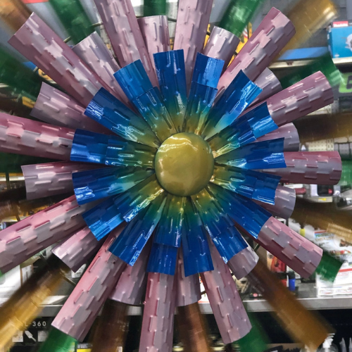 A vibrant, metallic flower sculpture with layered petals in blue, pink, and green, resembling a sunflower on display in a store setting.