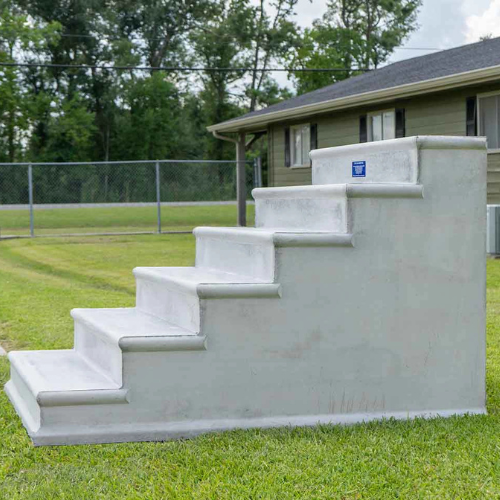 A large, white concrete storm shelter designed as outdoor steps is situated on a green lawn. A house and trees are visible in the background.