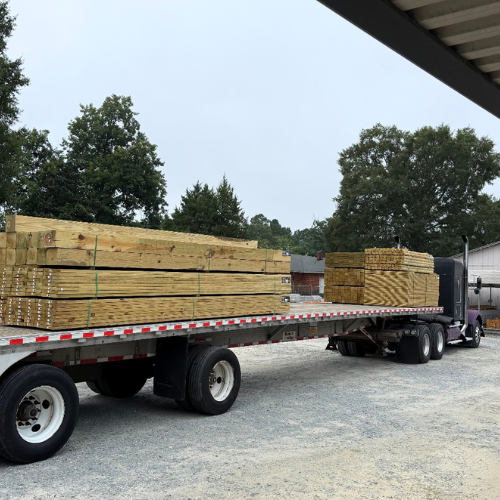 A flatbed truck loaded with neatly stacked lumber stands on a gravel lot. Tall trees and a clear sky are in the background. The mood is industrious.