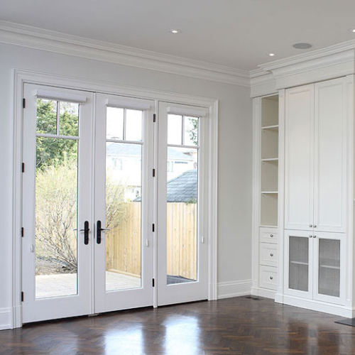 Bright room with glossy wooden floor, tall white French doors leading outside, and built-in white cabinets on the right. The mood is airy and elegant.