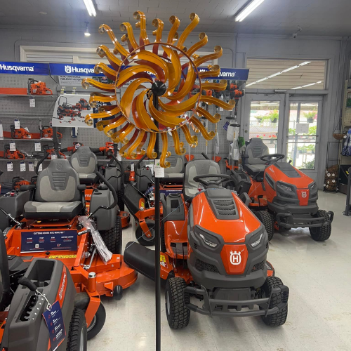 A display of orange and grey Husqvarna riding lawn mowers inside a store. A yellow and orange swirl sculpture hangs above them, creating a vibrant contrast.