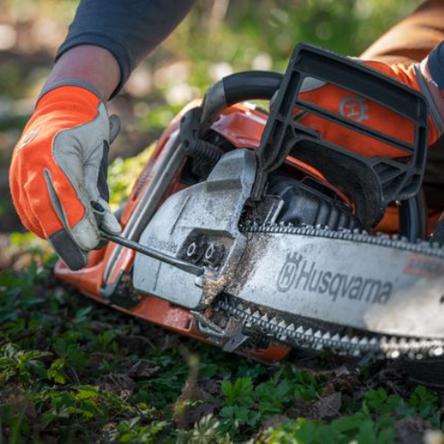 A person wearing orange gloves uses a wrench to adjust a Husqvarna chainsaw, set on green grass, conveying a focus on maintenance and safety.