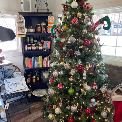 A festive Christmas tree adorned with red, silver, and green ornaments stands beside a bookshelf with jars and cookbooks in a cozy, window-lit room.