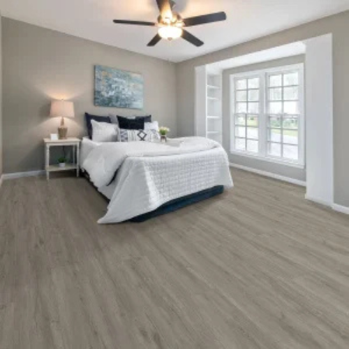Bright bedroom with gray hardwood floors and taupe walls. Features a bed with white bedding, bedside lamp, abstract wall art, and a bay window. Cozy and inviting.
