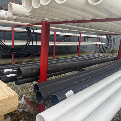 Rows of stacked white and black plastic pipes on red metal racks outdoors. Coiled cables are visible in the background, creating an industrial scene.