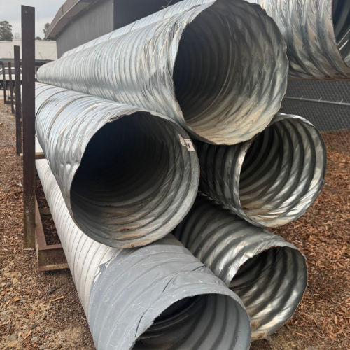 Large, stacked corrugated metal pipes are displayed outdoors on a wooden rack. The setting is industrial, and the light is overcast, creating a neutral tone.