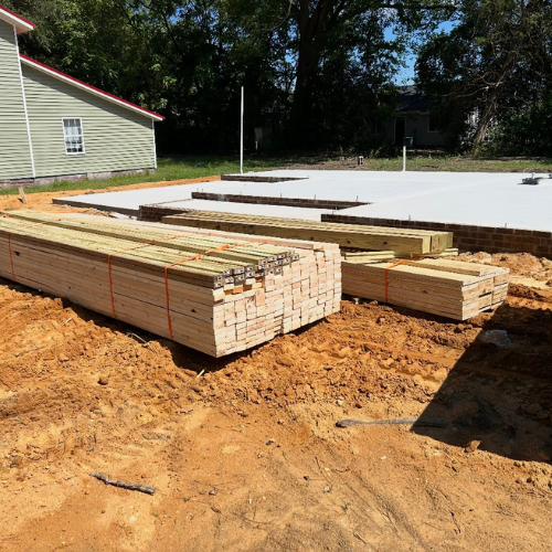 Stacks of lumber are neatly arranged on sandy soil at a construction site, with concrete foundation slabs in the background and trees nearby, under a clear blue sky.
