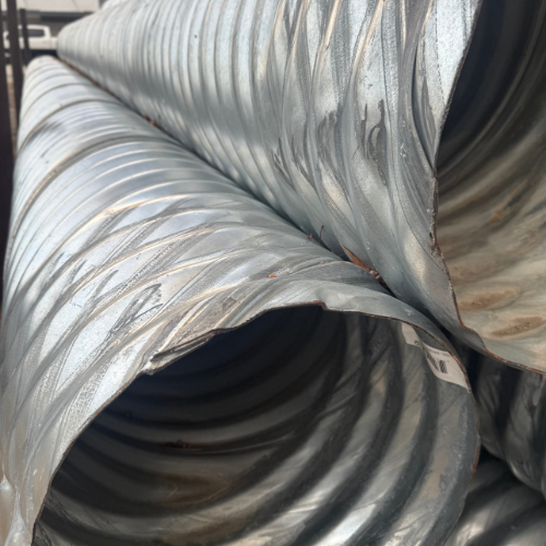 Stacked large metal pipes with ribbed surfaces in an industrial setting. The pipes, silver and cylindrical, create an orderly, textured pattern.