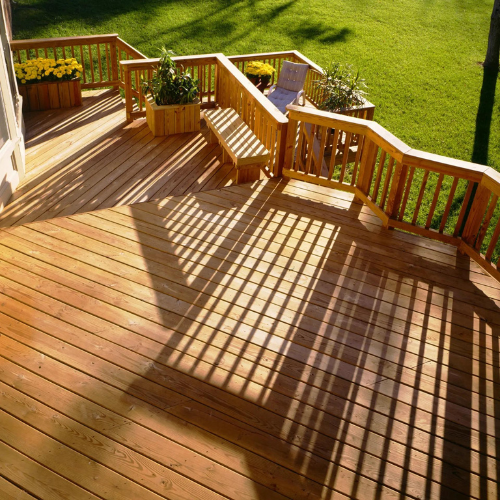Wooden deck with warm sunlight casting shadows through railings, leading to lush green lawn. Planters with yellow flowers and greenery add color.