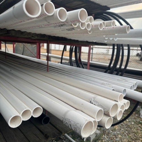 Close-up of stacked white PVC pipes on a shelf outdoors, with a metal chain-link fence in the background. The scene conveys an industrial and utilitarian tone.