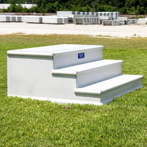 White three-step concrete block on green grass with trees and stacked concrete items in the background. The scene is bright and sunny.