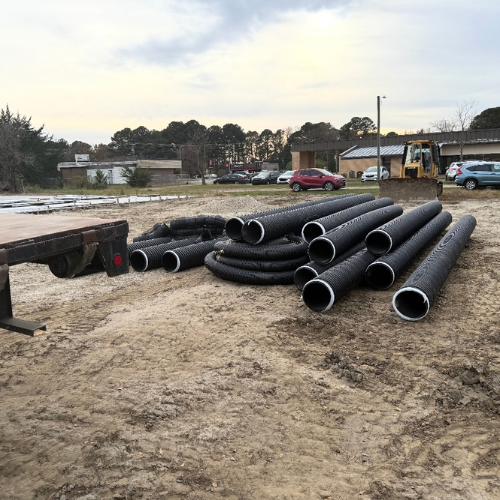 A construction site with large black pipes stacked on muddy ground, under a cloudy sky. Vehicles and trees are visible in the background.