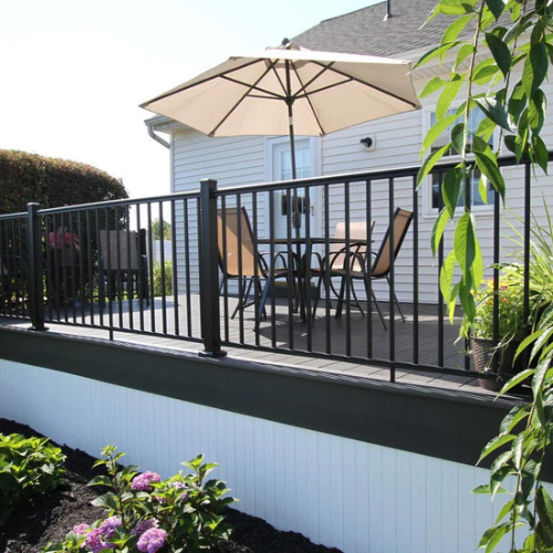 A sunny backyard with a wooden deck features a patio table, beige umbrella, and chairs. Surrounded by railings, greenery, and blooming flowers below.