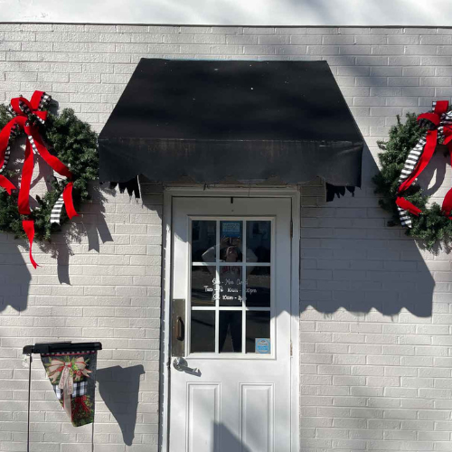 A white brick wall with a black awning shades a glass door. Two festive wreaths with red bows hang beside it, casting shadows in bright sunlight.