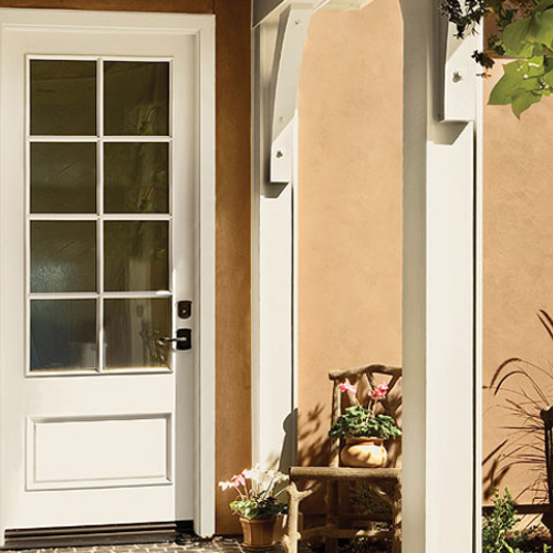 White door with glass panes next to a peach wall. Under a shaded porch, a wooden chair holds a pot of pink flowers, creating a serene, inviting atmosphere.