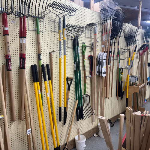 A variety of garden tools, including rakes and shovels with wooden and colored handles, neatly hang on a pegboard in a store setting.
