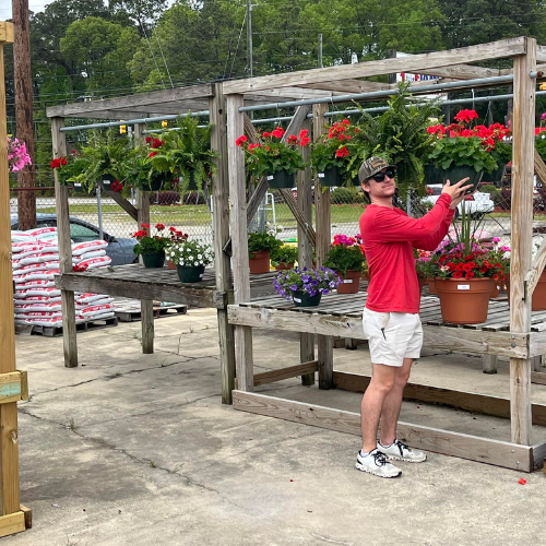A person in a red shirt and shorts, wearing a cap, poses cheerfully next to vibrant potted flowers on a wooden stand in an outdoor garden center.
