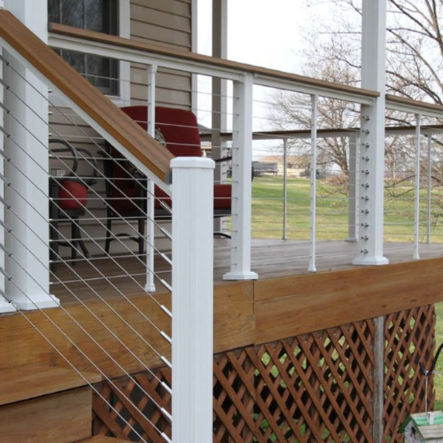 A wooden porch features a modern cable railing with white posts and a natural wood handrail. Red chairs are in the background, overlooking a grassy field.