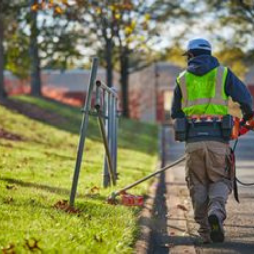 A worker in a high-visibility vest and helmet trims grass along a sidewalk using a string trimmer. The sun casts soft light and autumn leaves scatter around.