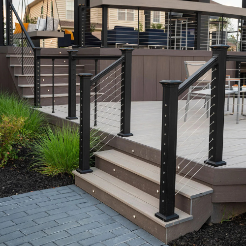 Modern deck with cable railing and steps leading to gray brick patio. Grassy plants are beside the stairs, creating a sleek and inviting outdoor space.
