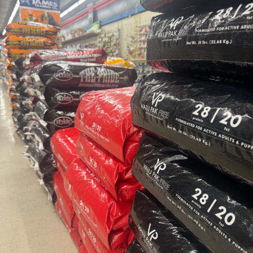 Stacks of red and black bags of dog food line a grocery aisle, creating a colorful, orderly display. Shelves in the background add depth and variety.