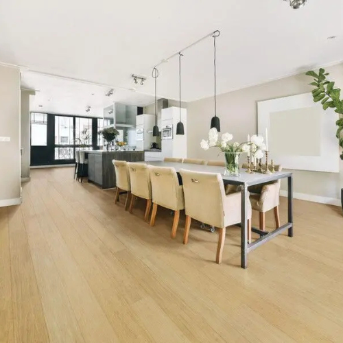 Spacious modern kitchen and dining area with light wood floors. Features a long dining table with beige chairs, hanging lights, and white cabinets.