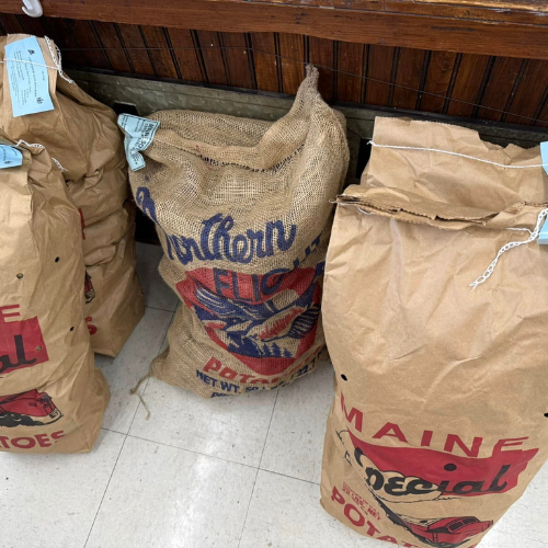 Three large bags of potatoes, one burlap and two brown paper, each labeled with different brands, rest on a tile floor against a wooden wall.