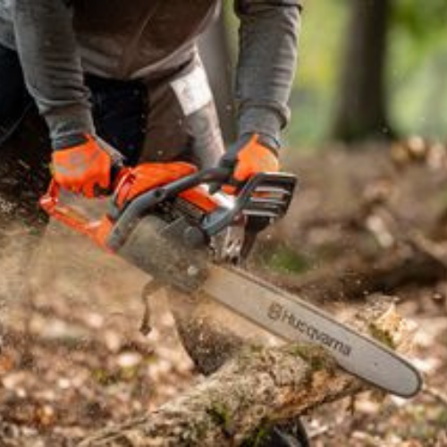 A person in gray clothes and orange gloves uses a Husqvarna chainsaw to cut a tree branch. Wood chips fly, creating a dynamic and focused tone.