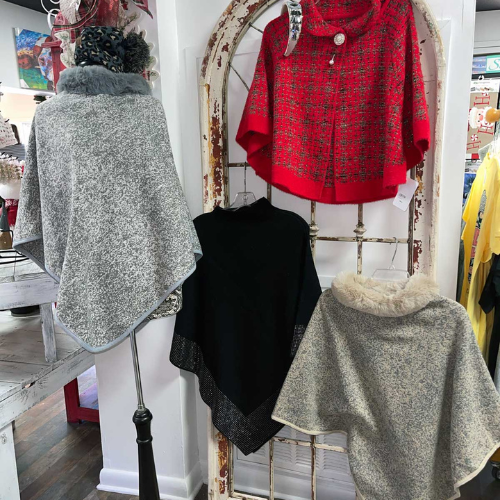 A display of elegant ponchos and capes: a gray cape with fur trim, a black poncho with a sparkly edge, and a vibrant red, patterned cape. Warm, cozy ambiance.