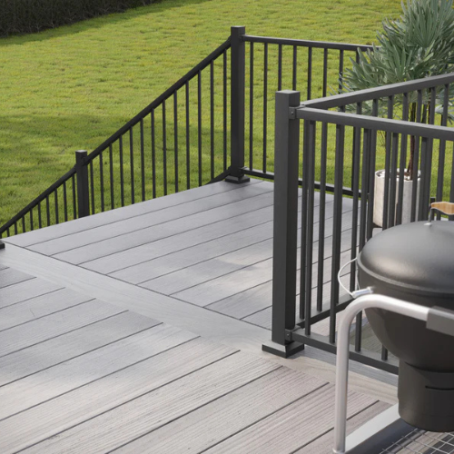 Modern outdoor patio with gray composite decking and black metal railings, overlooking a lush green lawn. A black barbecue grill is visible in the foreground.