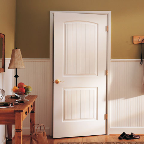 A white interior door with decorative panels is ajar in a warm, cozy room. A wooden table with a fruit bowl and lamp is nearby; soft lighting casts gentle shadows.