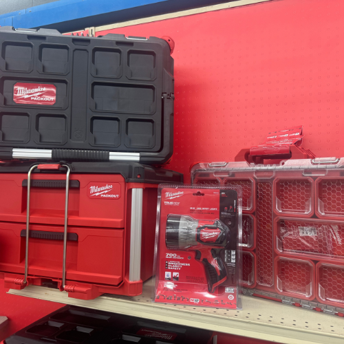 A store shelf displaying Milwaukee Packout toolboxes and a cordless drill set. The red and black color scheme conveys a professional, rugged tone.