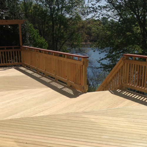 A spacious wooden deck with railing overlooks a tranquil lake surrounded by lush trees. The sunlight casts soft shadows, creating a serene and inviting atmosphere.