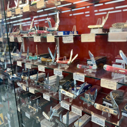 A display case filled with various pocket knives, each with open blades on glass shelves. Price tags are visible. Brightly lit with a red background.